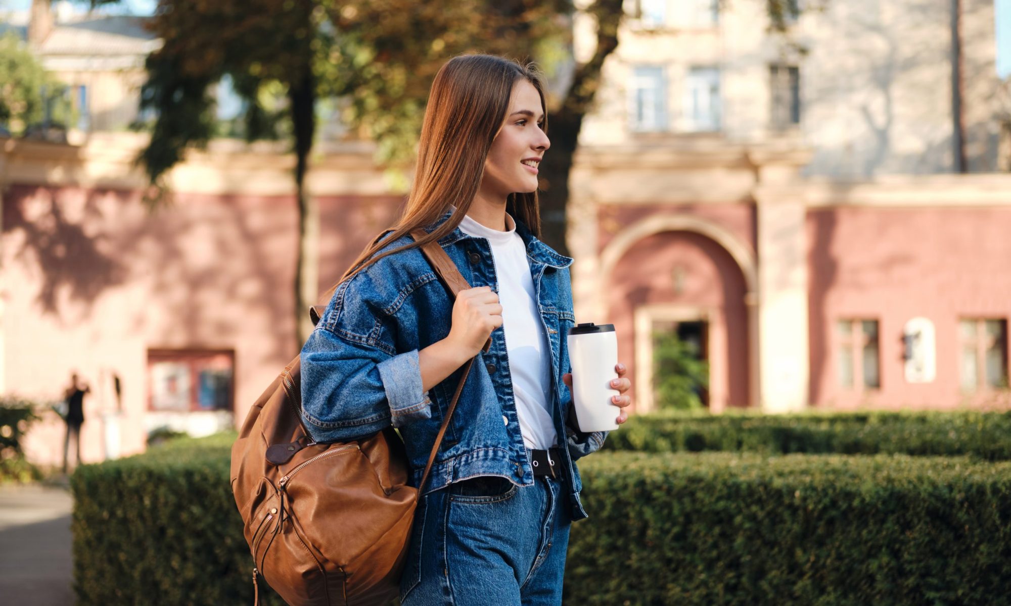 Side view of casual student girl in denim jacket with backpack and coffee happily looking away in college campus outdoor