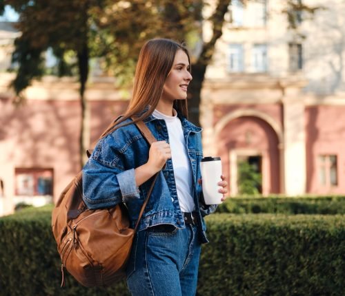Side view of casual student girl in denim jacket with backpack a Side view of casual student girl in denim jacket with backpack and coffee happily looking away in college campus outdoor